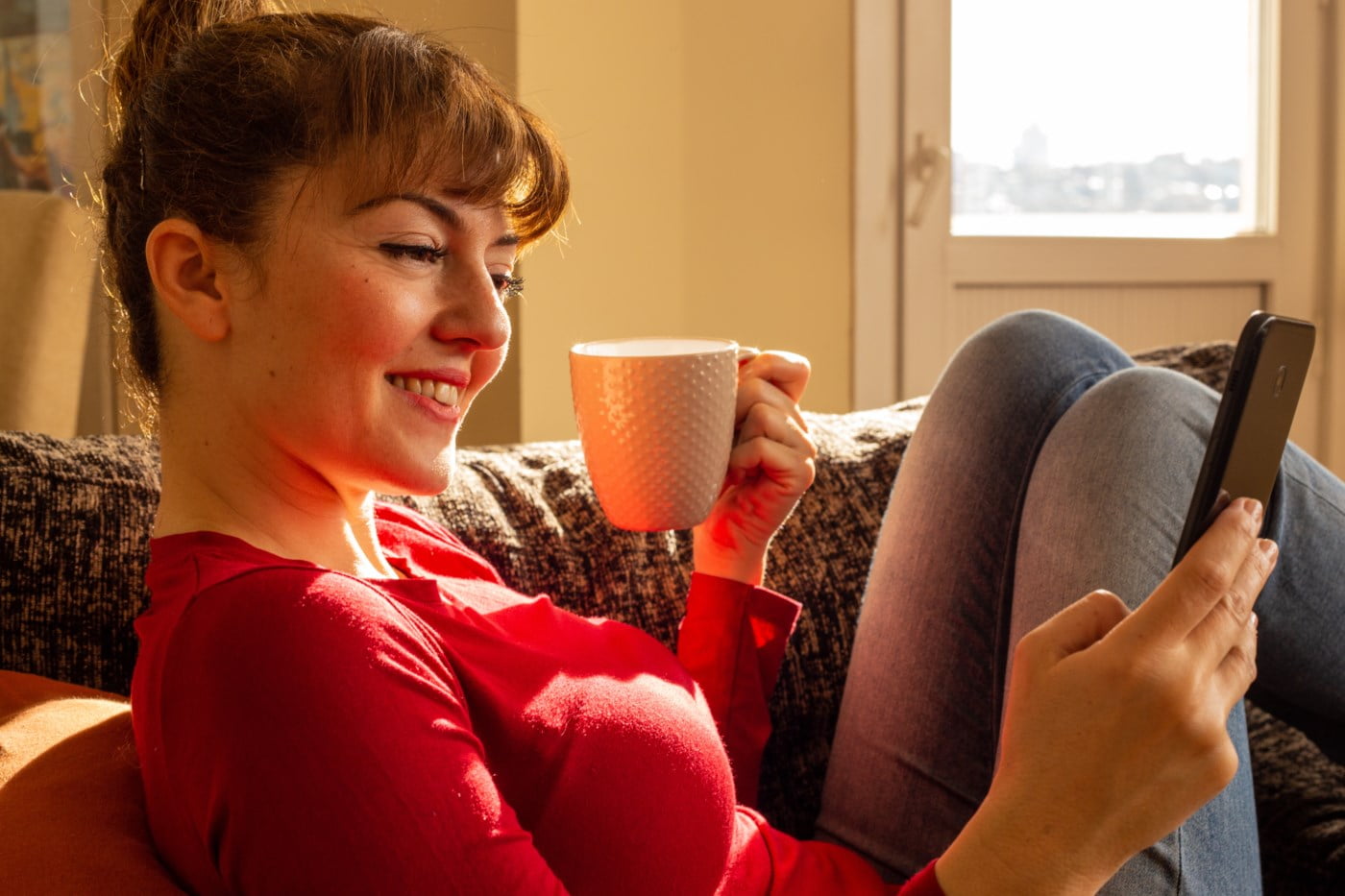 Kvinne smiler medan ho sit i sofaen med ein kaffikopp i handa og mobilen i fanget. Ho ser på skjermen i varmt dagslys.