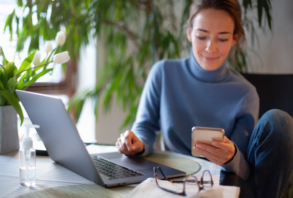 En kvinne sitter med pc-en sin foran seg i mens hun ser på telefonen sin