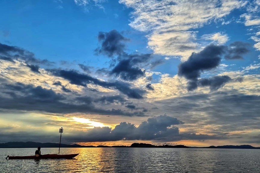 Person som ror i kano på sjøen når det er solnedgang og blå himmel med noen skyer på